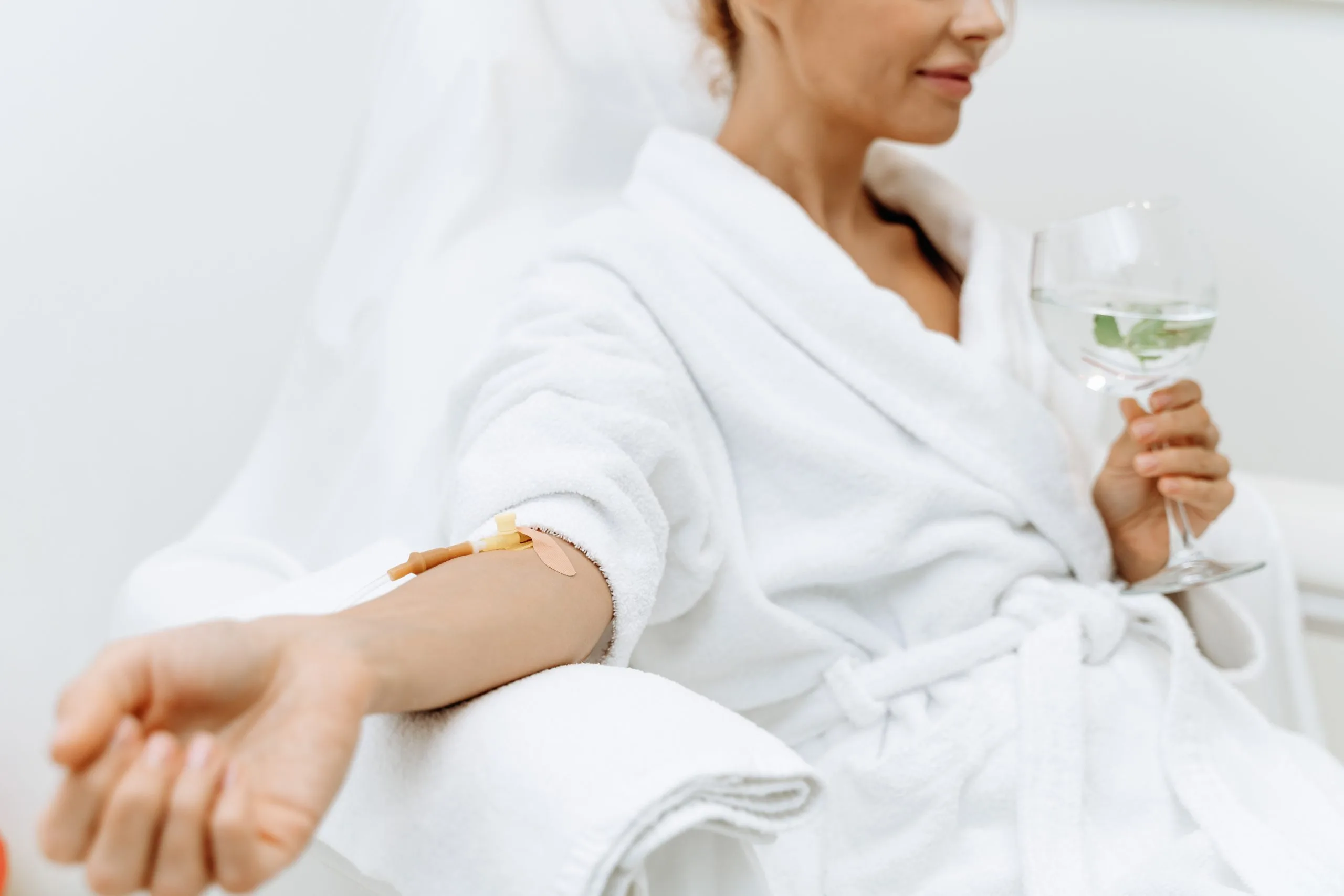 Woman in white robe holding water glass with bandage on inner elbow after treatment