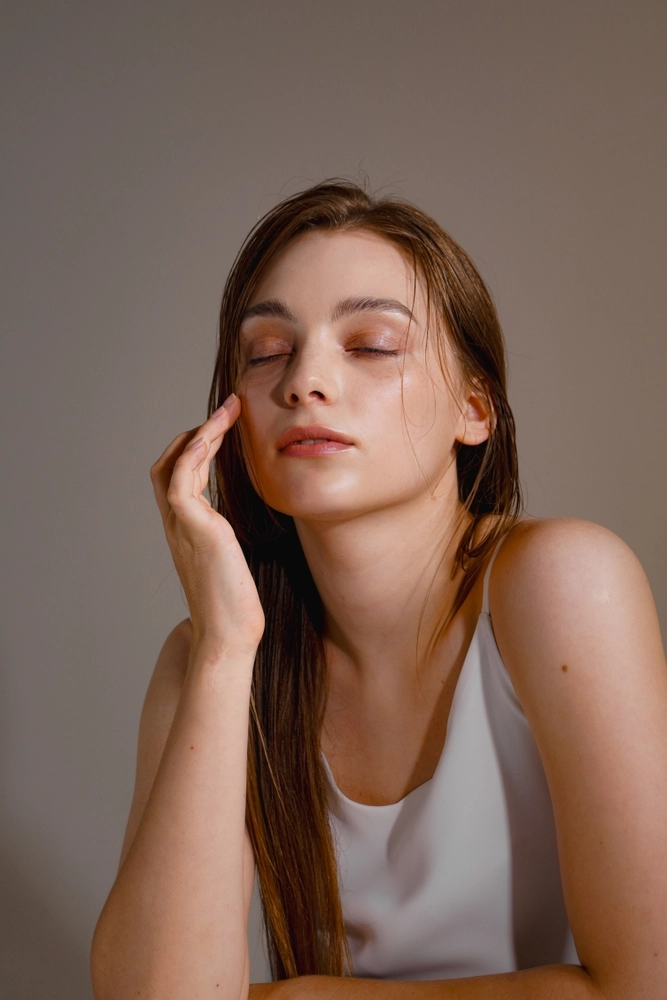 Woman with closed eyes touching her cheek with hand against neutral background