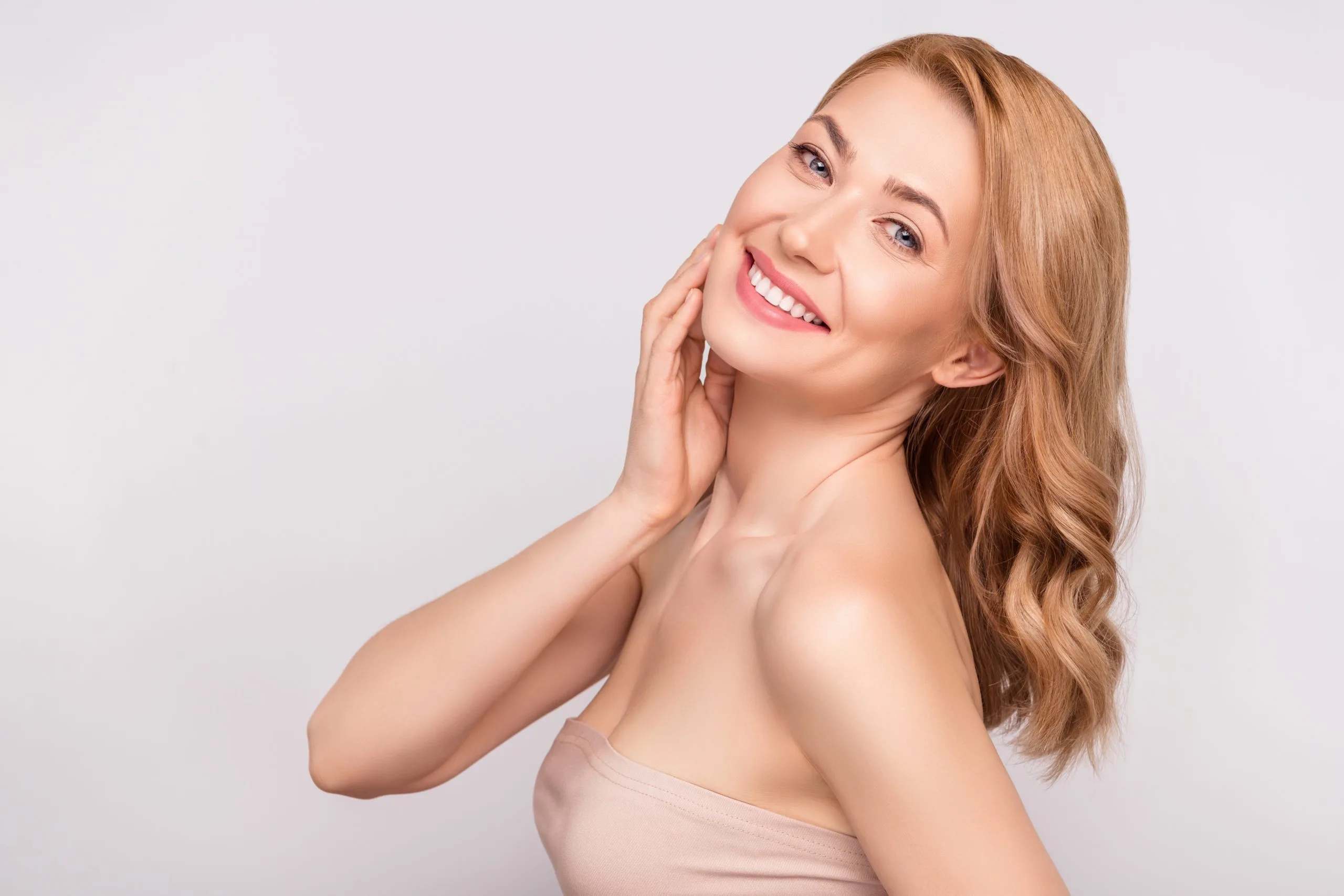 Woman with brown wavy hair smiling and touching her face against a light gray background