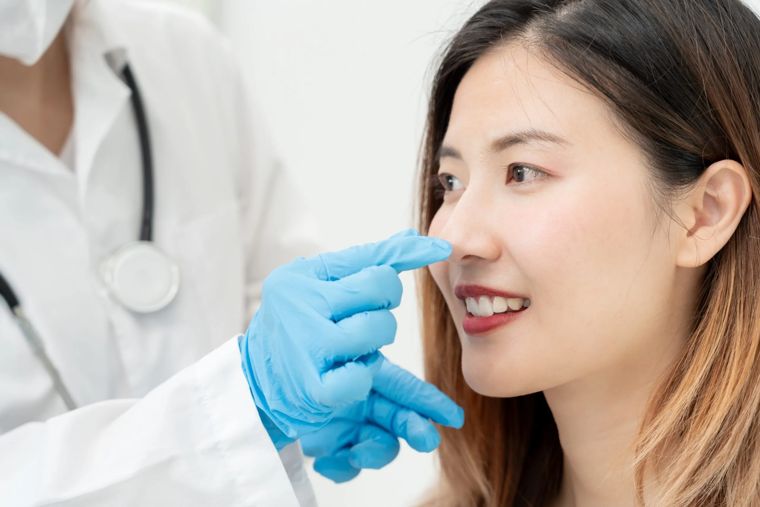 Doctor in white coat and blue gloves pointing to patient's nose during cosmetic consultation